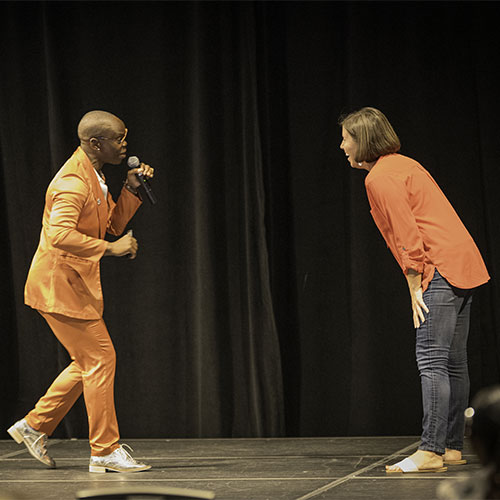 A person in an orange suit leans forward holding a microphone toward an audience member on a stage