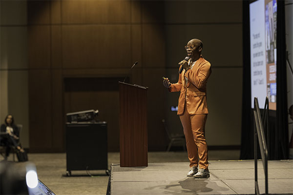 A person in an orange suit speaks on stage at an event, holding a microphone.